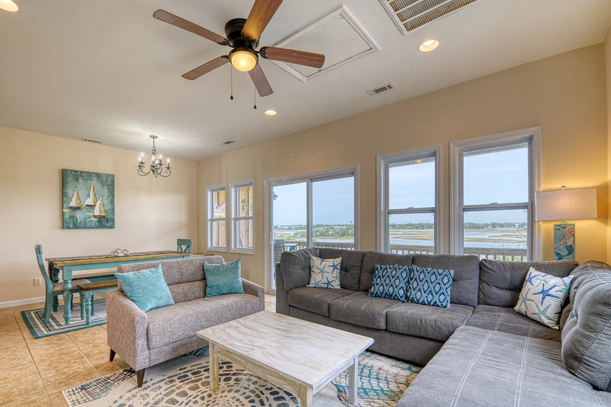 An open-plan living and dining area with a large sectional sofa, a spacious dining table with chairs, and floor-to-ceiling windows that flood the room with natural light. Decorative wall art adds warmth, while the windows showcase scenic water views. The inviting arrangement encourages socializing, relaxation, and shared meals.