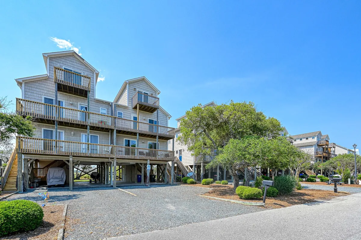 The image showcases a cheerful, three-story beach house in Surf City, North Carolina, with multiple balconies overlooking a landscaped yard that slopes down to the water. The sunset glows warmly over spacious decks perfect for relaxing and gathering, inviting outdoor barbecues and morning coffee with picturesque marsh views.