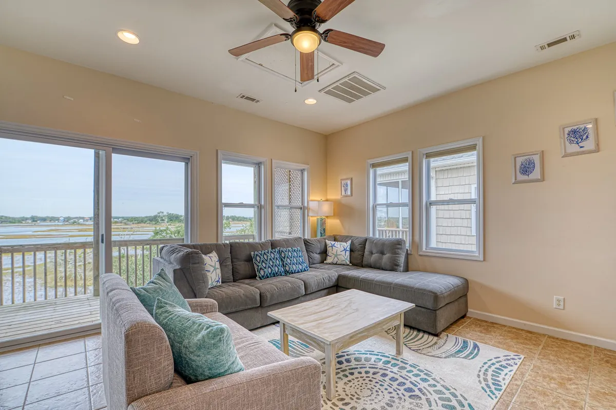 A spacious living area with a large sectional sofa arranged around a coffee table, with windows letting in natural light and providing a relaxing view. Decorative wall art enhances the cozy ambiance, making it ideal for gatherings or unwinding after a day at the coast.