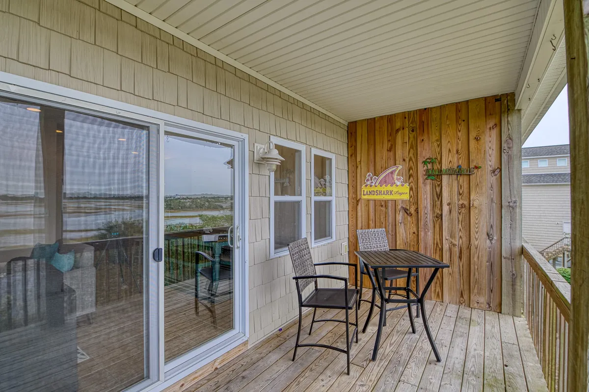 An exterior shot of a deck with a bistro table and chairs, wooden wall paneling, and a water view visible through a sliding door. The setting suggests a relaxed space for enjoying coffee, casual conversation, or simply savoring the view.