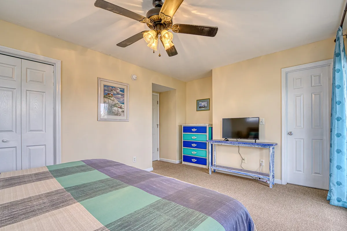 A cozy bedroom featuring a bed with colorful bedding, a ceiling fan with light fixture, a blue dresser, and a television on a stand. Natural light fills the space, creating a welcoming atmosphere for restful nights or private retreats.