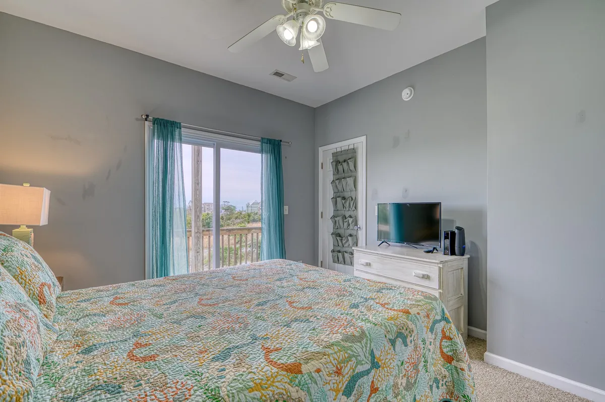 A comfortable, serene bedroom with a bed adorned in a colorful quilt, sheer curtains filtering soft light through the window, and a TV atop a dresser. The room exudes calm and offers a retreat for relaxation either during the day or in the evening.
