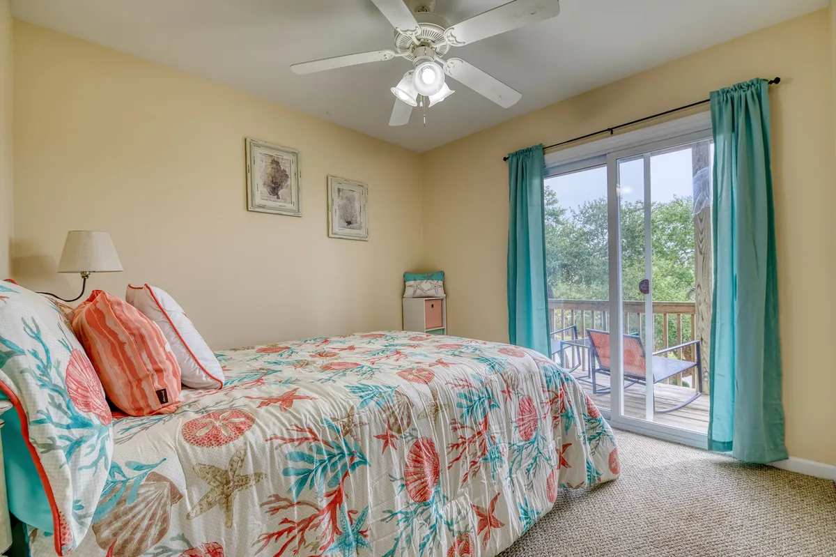 A tranquil guest room showcasing a bed with a colorful quilt, a sliding glass door that opens out to a private deck, and tasteful wall art. Natural light fills the space, and the deck offers a glimpse of peaceful greenery beyond, inviting relaxation and quiet moments.